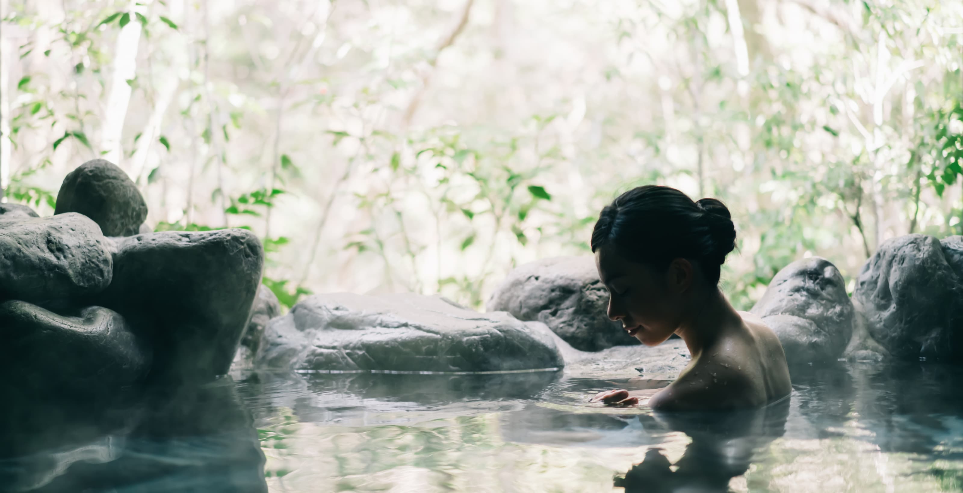 A person relaxing in a secluded hot spring surrounded by natural rocks and dense green foliage, evoking a sense of calm and connection with nature.