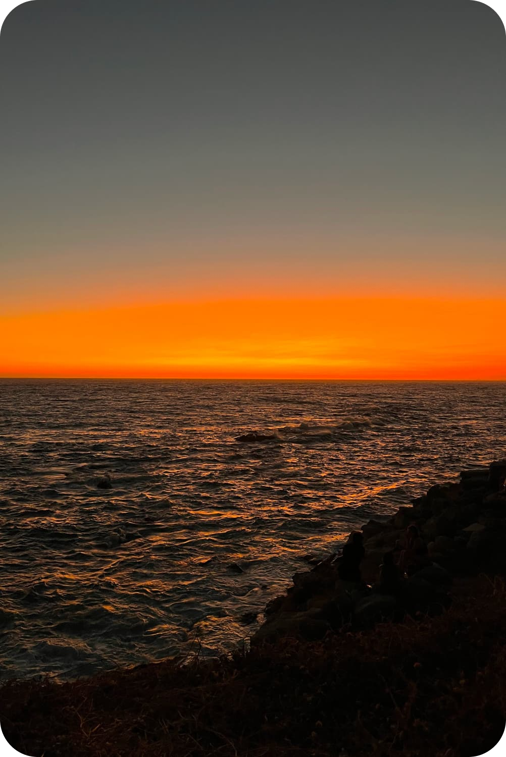 A captivating sunset over the ocean with a gradient of fiery orange to deep blue colors in the sky, and silhouettes of people on rocky shores observing the tranquil scene.