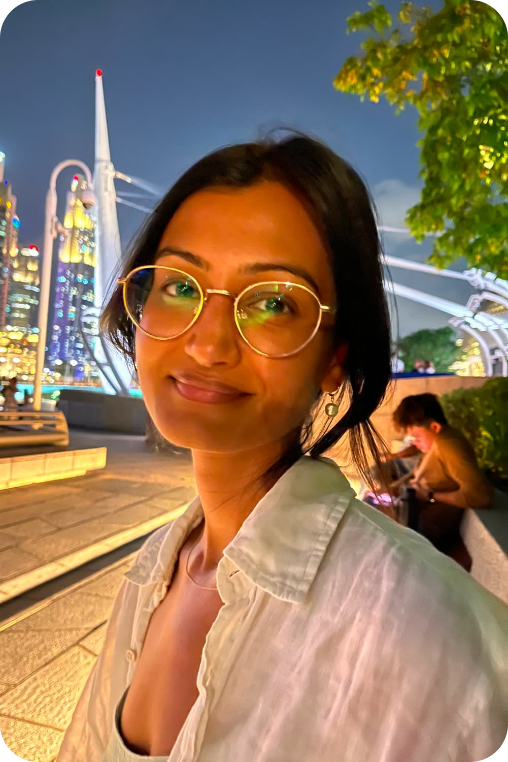 A close-up smiling picture of Oshadi's founder Ritu, captured at night with the vibrant, illuminated cityscape of Singapore and architectural structures in the background.