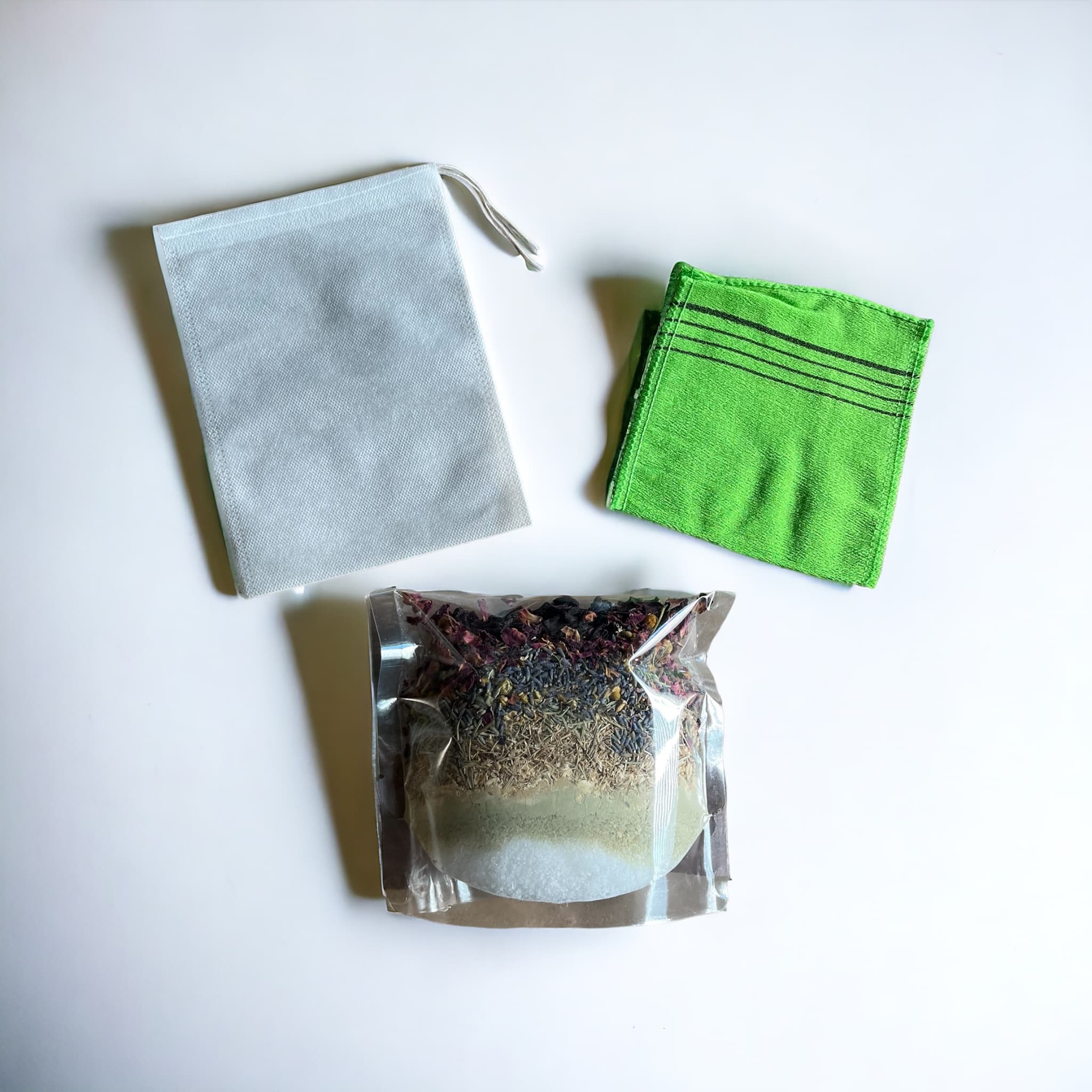 A transparent plastic package filled with a layered mix of dried herbs, salts, and flowers for a bath blend, next to a large muslic bath tea bag and a green exfoliating cloth on a white surface.