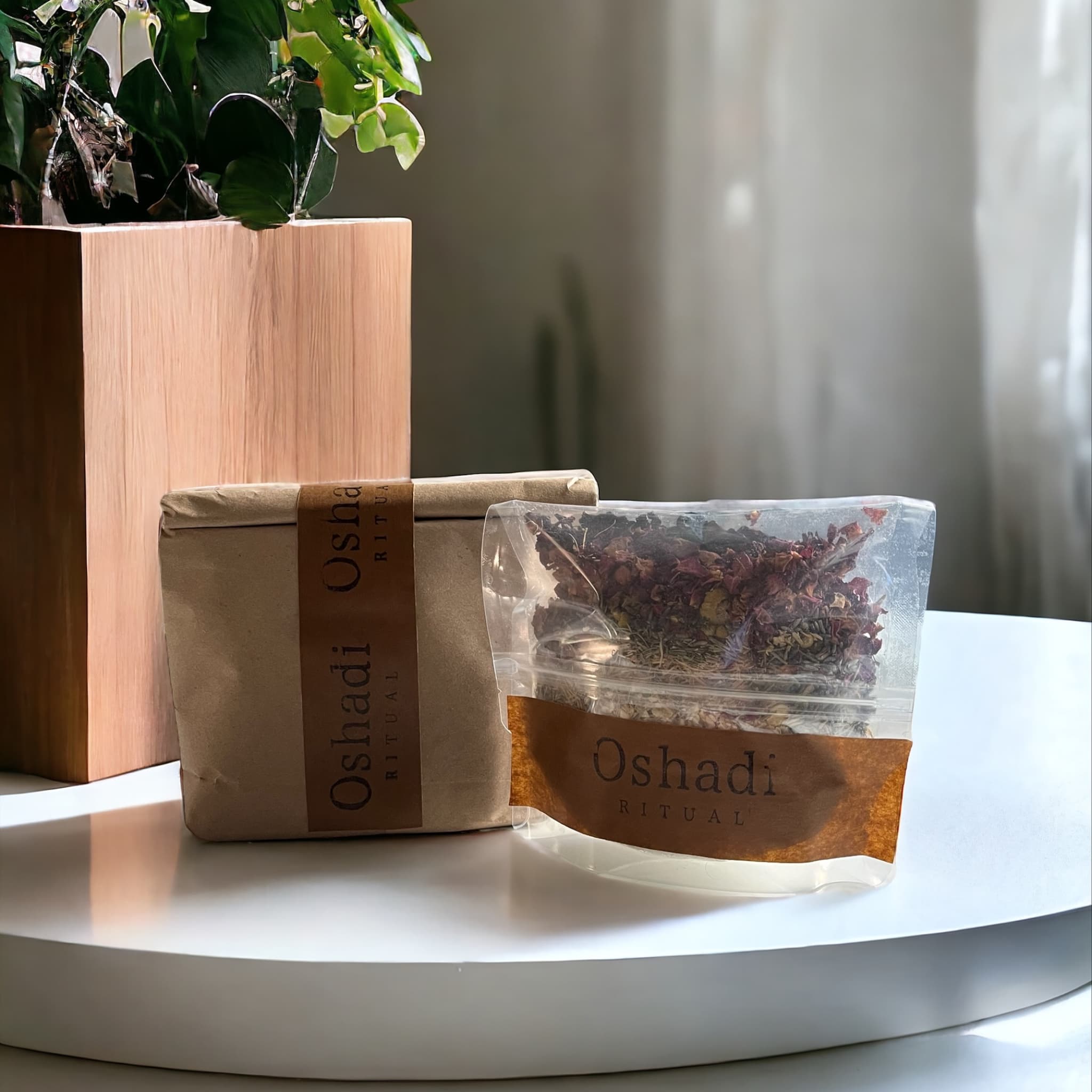 Sustainable brand packaging for Oshadi Ritual, featuring a clear plastic bag with layered bath blend contents beside its minimalist brown paper packaging on a wooden table.