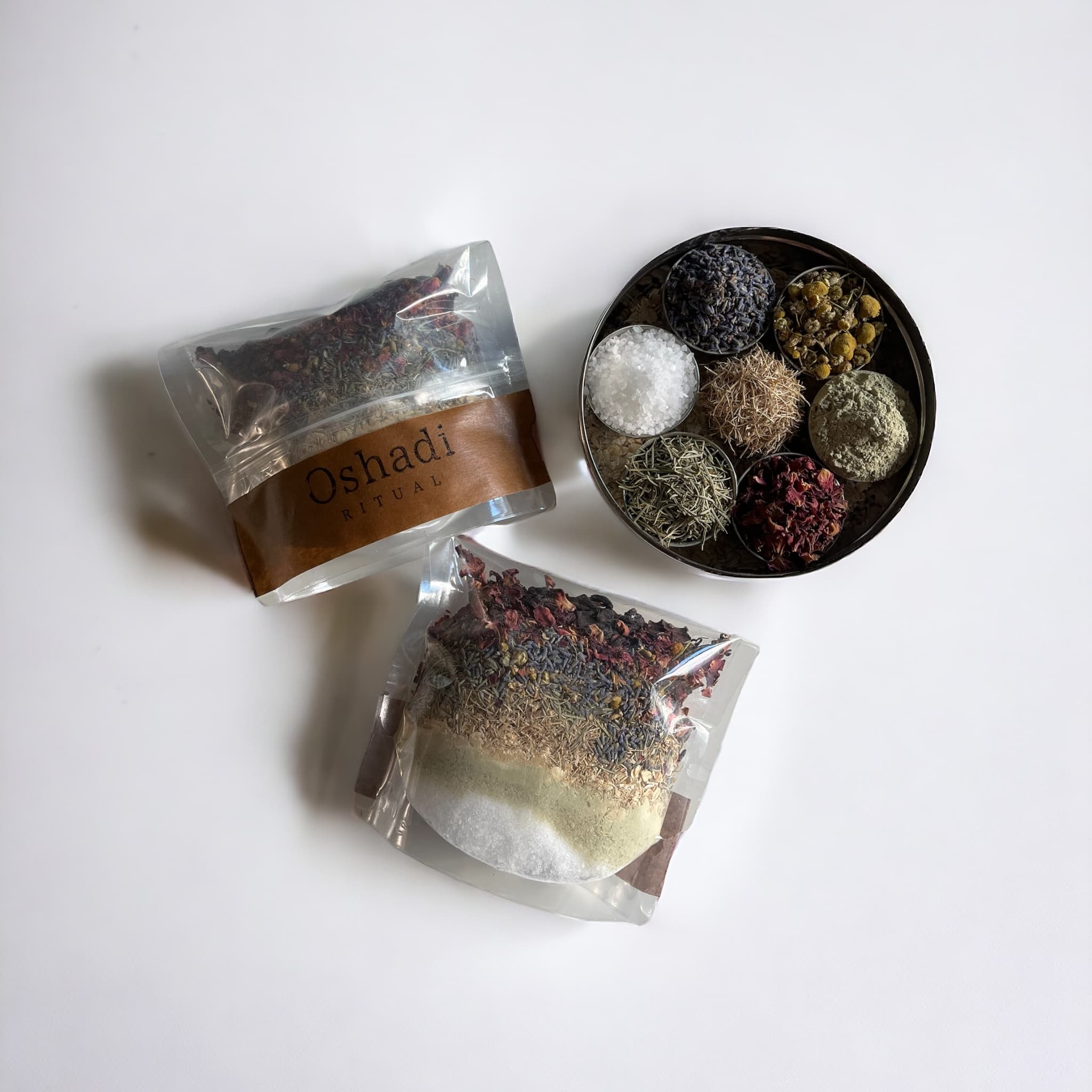 A clear plastic bag branded with 'Oshadi Ritual' containing a colorful bath blend displayed alongside an open metal tray with neatly arranged ingredients like lavender, chamomile, and salts.