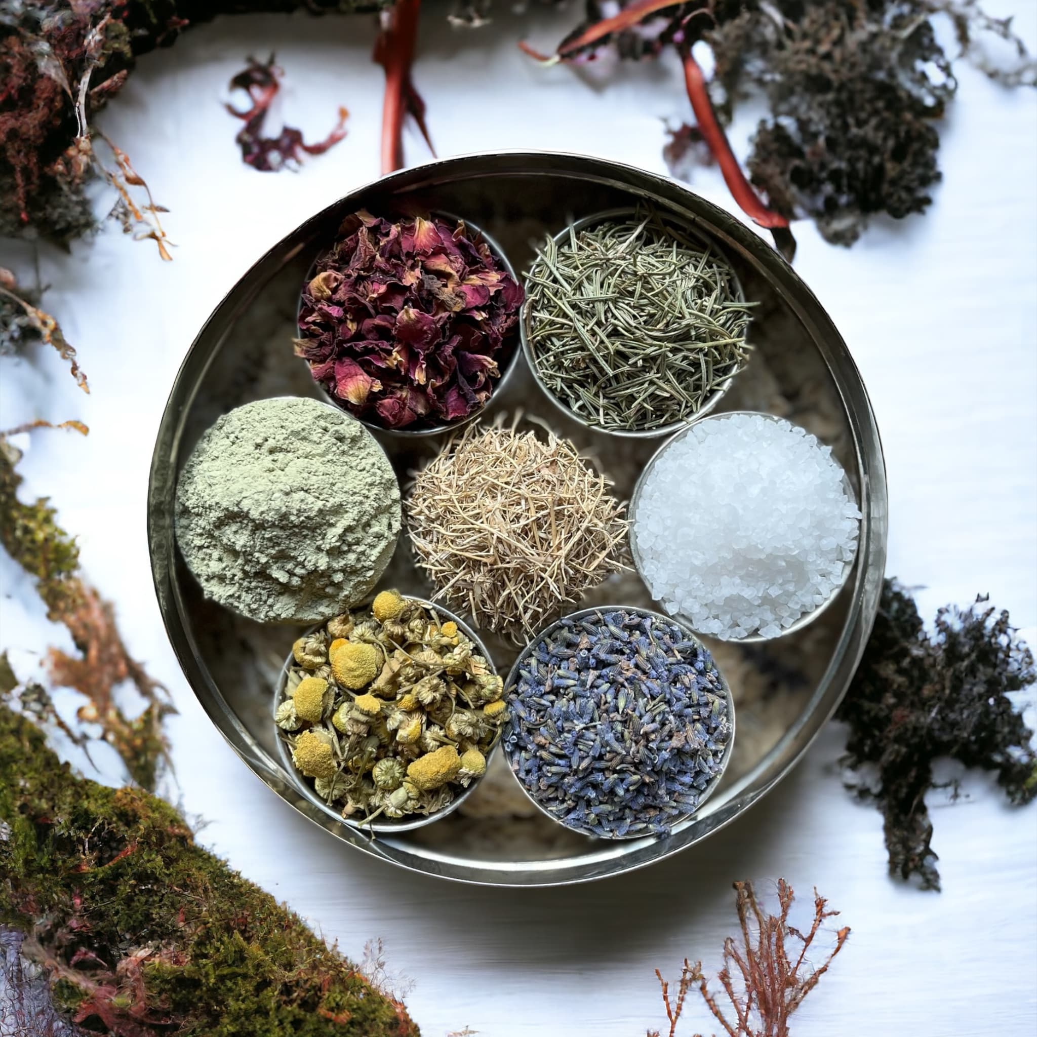 A top-down view of a circular tray divided into sections containing various dried herbs and minerals, including rose petals, chamomile, and coarse sea salt, set against a backdrop of assorted dried plants.