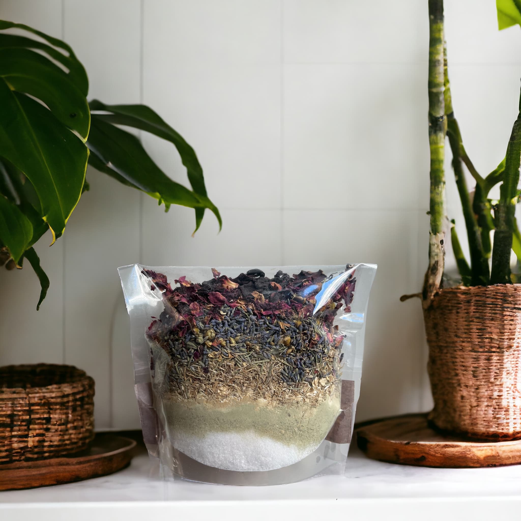 A clear, resealable plastic bag on a bathroom counter filled with layered bath blend ingredients including dark red rose petals, blue lavender buds, and beige botanical powders.